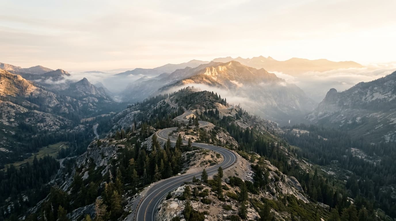 A wide aerial of a switchback road climbing a misty ridge toward bright light.